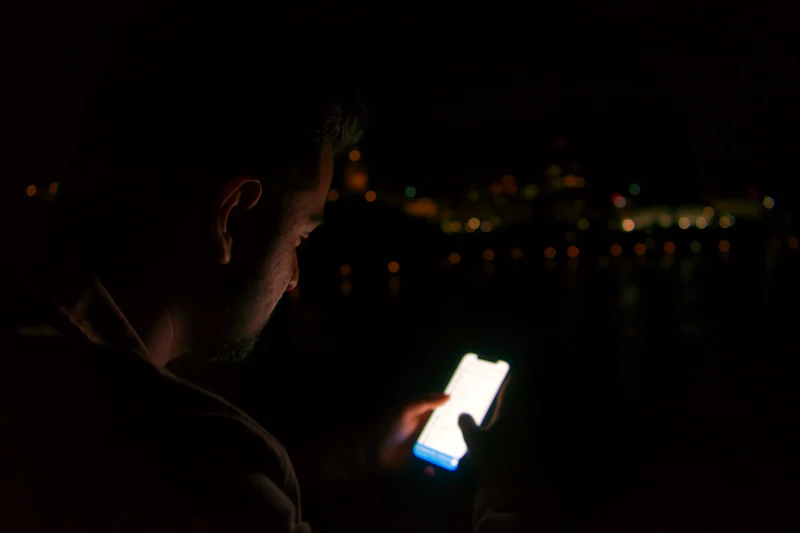 A man in a dark room, his face lit by his phone screen.