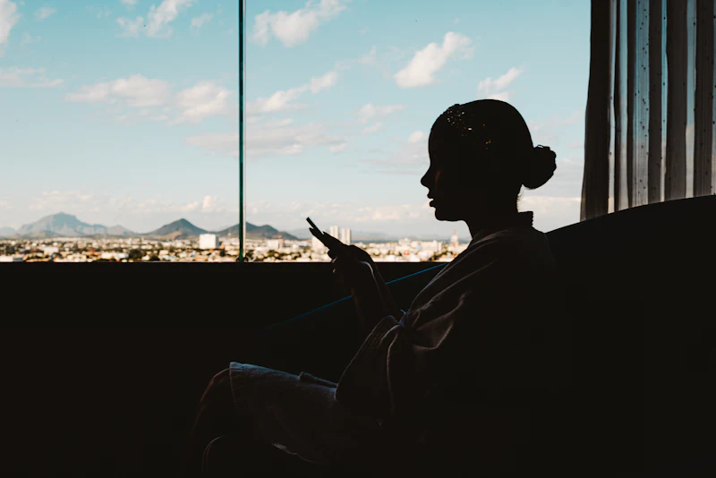 Silhouette of a woman checking her phone by a window, blue sky outside.